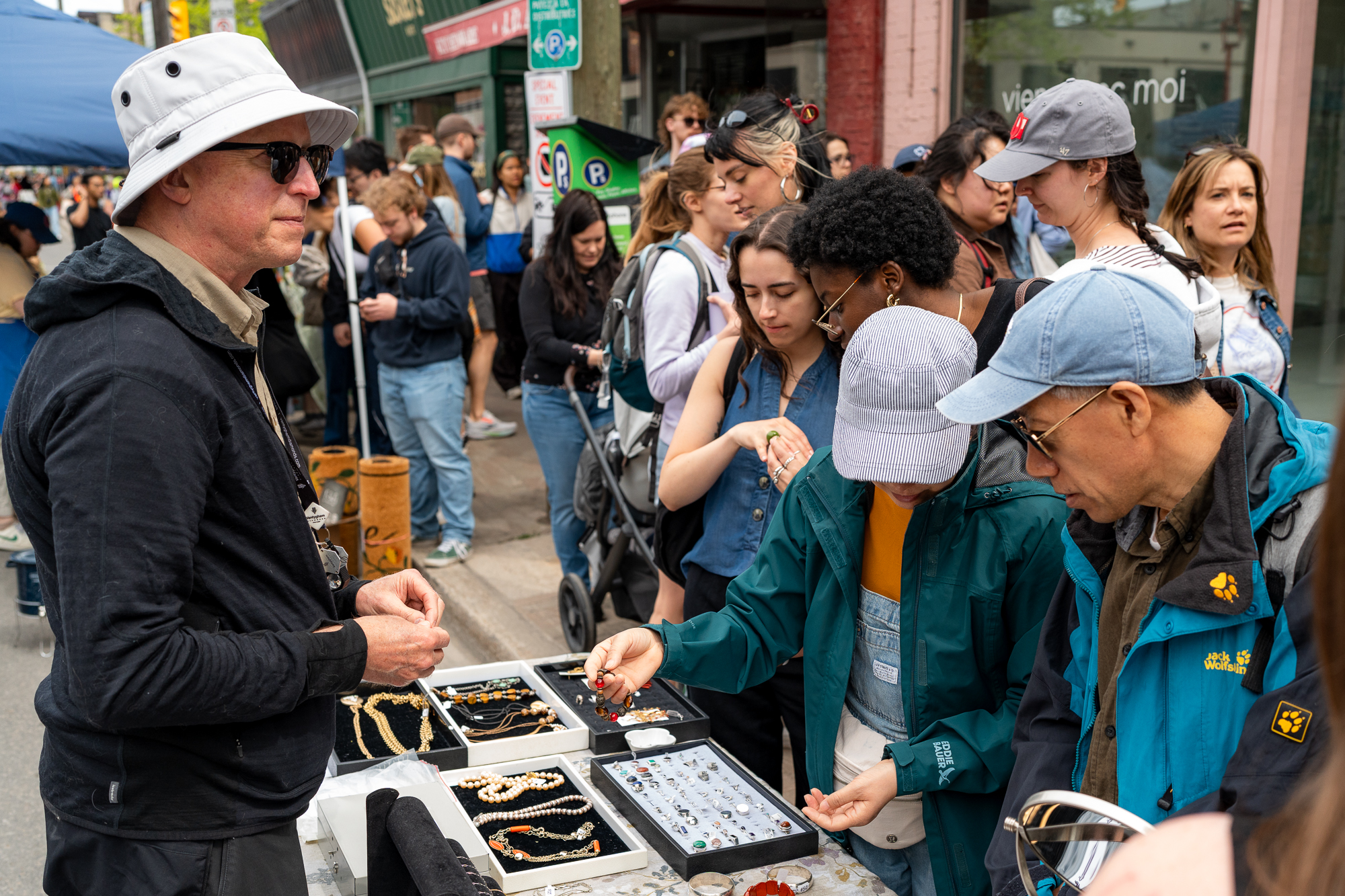 A vendor table of rings and necklaces drawing a sidewalk crowd on Bank Street in midsummer